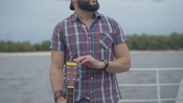 Oigenkännlig man som står med gitarr på flodstranden och tänker. Ung kaukasisk ensam musiker tillbringar molnig sommardag på vall med stränginstrument. — Stockvideo