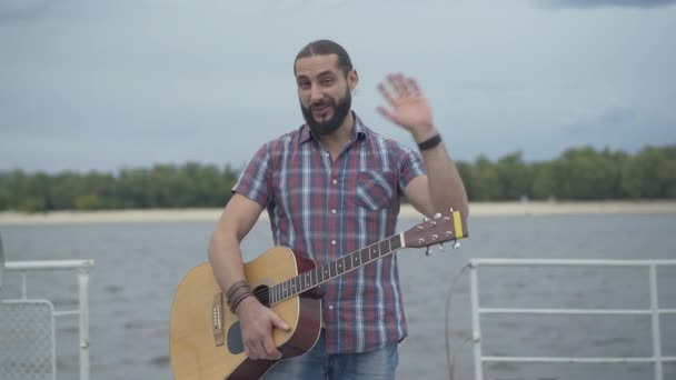 Positive Caucasian man standing with guitar on embankment and waving. Portrait of happy young handsome musician looking at camera and smiling. Joy and happiness concept. — Stock Video