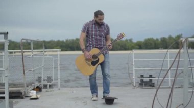 Nehir kıyısında setin üzerinde duran kahverengi şapkalı sokak müzisyeninin geniş açılı görüntüsü. Konsantre gitarist para kazanmak için açık havada müzik aleti çalıyor. Dilenci konsepti.