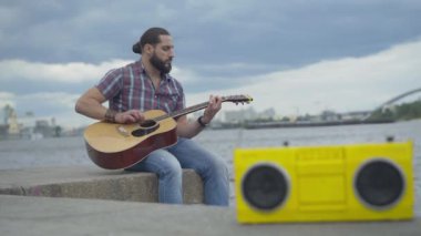 Önünde bulanık sarı bant kayıt cihazıyla şehir setinde gitar çalan genç bir müzisyen. Nehir kıyısında sahne alan odaklı beyaz adamın portresi. Sanat ve yaşam tarzı.