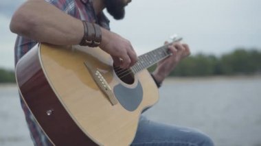 Gitarın yakın çekimleri beyaz erkeklerin elinde. Tanımlanamayan genç adam arka planda gri bulutlu gökyüzü ve nehir ile telli müzik aleti çalıyor..