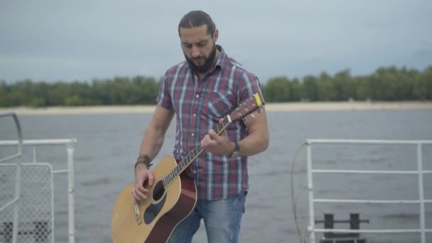Уличный музыкант, играющий на гитаре на набережной, как неузнаваемая женщина, проходящая впереди. Молодой белый мужчина присматривает за дамой. Бородатый гитарист выступает на берегу реки. — стоковое видео
