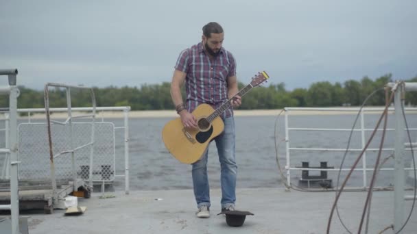 Wide shot of absorbed street musician performing on river bank with brown hat lying on embankment. Concentrated guitarist playing musical instrument outdoors to earn money. Beggary concept. — Stock Video