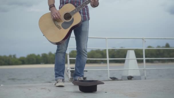 Benet på en oigenkännlig gitarrist som träffar en brun hatt som ligger på vallen. Arg olycklig vit man som spelar musikinstrument på flodstranden. Fattigdom och tiggeri. — Stockvideo