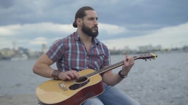 Brunette Caucasian bebaarde man speelt gitaar met wazig stad op de achtergrond. Zelfverzekerde jonge muzikant geniet van hobby op stedelijke dijk. Hobby en levensstijl. — Stockvideo