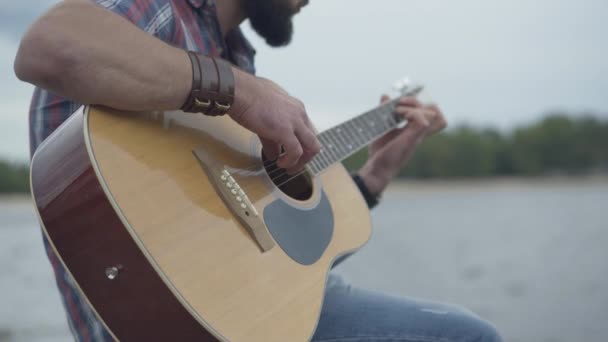 Gitarın yakın çekimleri beyaz erkeklerin elinde. Tanımlanamayan genç adam arka planda gri bulutlu gökyüzü ve nehir ile telli müzik aleti çalıyor.. — Stok video