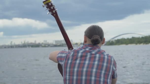 Widok z tyłu białego człowieka siedzącego z gitarą na brzegu rzeki z zamazanym miastem w tle. Przemyślany młody gitarzysta spędzający samotny dzień na świeżym powietrzu. — Wideo stockowe
