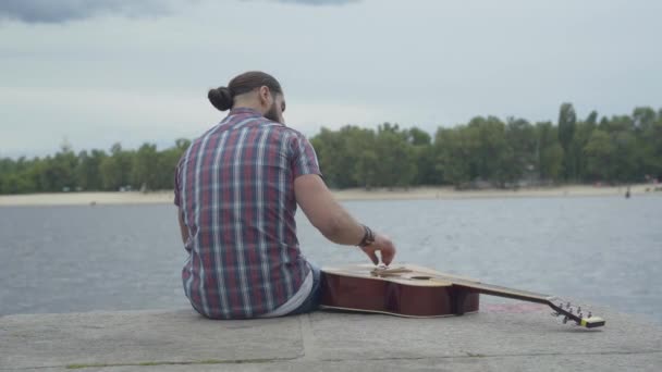 Back view of frustrated man sitting with guitar on river bank and throwing stones in water. Depressed Caucasian young musician spending lonely cloudy summer day outdoors. — Stock Video