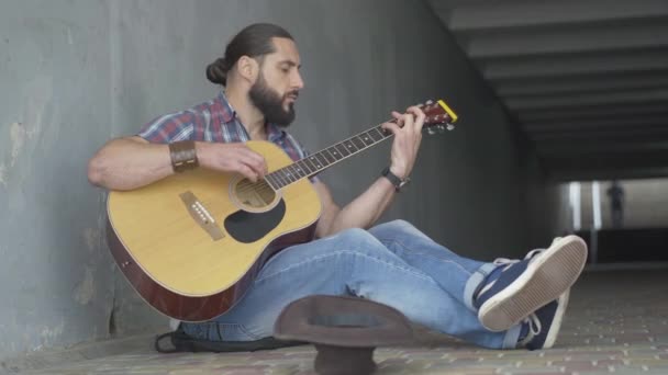 Ung manlig tiggare som spelar gitarr i urban underjordisk korsning. Porträtt av olycklig fattig vit man sittandes med musikinstrument och hatt. Street musik i staden. — Stockvideo