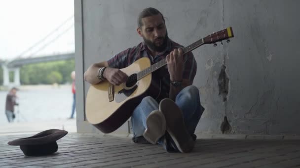 Porträtt av dålig musiker som spelar gitarr i underjordisk korsning vid flodbanken. Sorgligt skäggig vit man som uppträder på stränginstrument utomhus. — Stockvideo