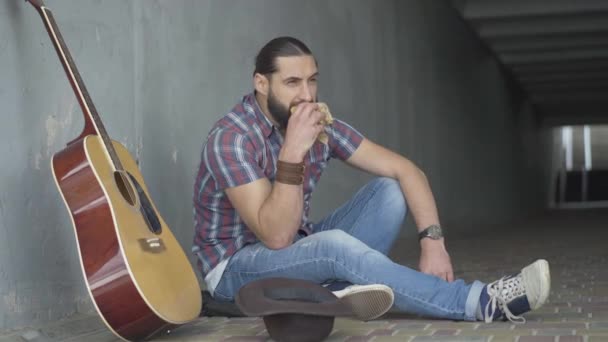 Poor Caucasian man sitting with guitar and hat in urban underground crossing and eating bun. Wide shot portrait of male musician having break performing outdoors in city. — Stock Video