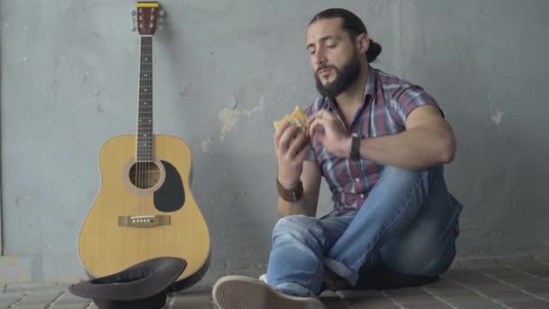 Brett skott framifrån av ung frustrerad man sitter i underjordisk korsning med gitarr och äta. Porträtt av vit musiker som äter lunch efter tiggeri. — Stockvideo