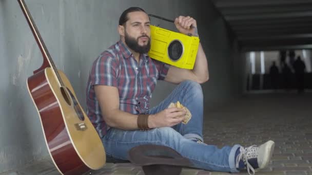 Lonely poor Caucasian musician eating bun and listening to music from retro tape recorder. Portrait of sad man sitting in underground crossing with guitar. Poverty and begging in modern society. — Stock Video