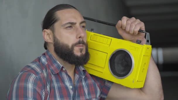 Close-up of young Caucasian man with brown eyes listening to music from vintage yellow tape recorder and eating bun. Portrait of poor street beggar having lunch in urban underground crossing. — Stock Video