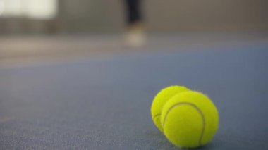 Spor salonunda yerde yatan sarı tenis toplarının yakın plan çekimleri ve arka planda bulanık beyaz kadın servis yapıyor. Yarışmadan önce iç sahada tanınmayan kadın antrenmanı.