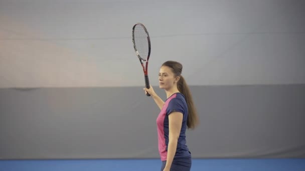 Camera nadert geconcentreerde blanke vrouw serveert tennisbal met racket. Zijaanzicht portret van zelfverzekerde sportvrouw start wedstrijd in de sportschool op indoor court. — Stockvideo