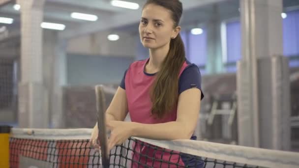 Mosolygó barna sportos nő pózol tenisz ütő a neten. Portré egy elbűvölő, fiatal kaukázusi nőről, aki tornateremben edz. Versenyképes sportkoncepció. — Stock videók