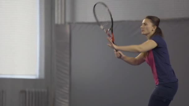 Live camera volgt beweging van geconcentreerde sportvrouw spelen grote tennis in de sportschool. Portret van een blanke vrouw die voor de wedstrijd binnenshuis traint. Workout van atletische jonge speler. — Stockvideo