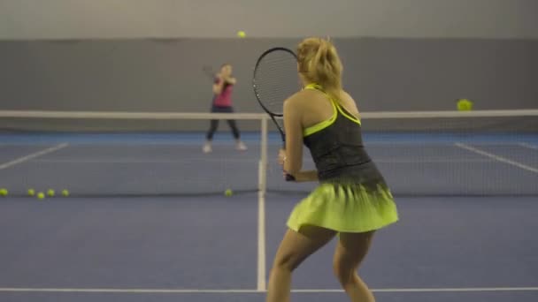Back view of young slim blond woman training in gym with blurred partner. Live camera follows movement of professional young Caucasian sportswoman hitting ball with racket indoors. — Stock Video