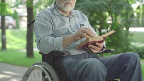 Homme âgé paralysé méconnaissable tournant la page du livre et lisant à haute voix. Vieux retraité caucasien aux cheveux gris profitant de la littérature dans un parc d'été ensoleillé. Hobby et handicap. — Video