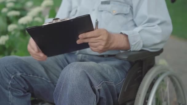 Nerozpoznatelný paraplegický starší muž podepisující venku dokumenty. Starý běloch paralyzoval důchodce, který seděl na vozíčku a psal podpis na papíry. Koncept sociálních služeb. — Stock video