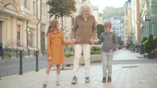 Bred bild av glada kaukasiska farfar promenader med söt pojke och flicka utomhus på solig sommardag. Glada gråhåriga äldre man och positiva barnbarn promenader längs gatan i staden. — Stockvideo