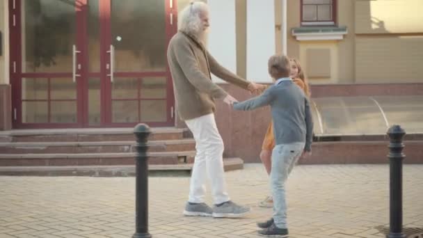 Caméra suit joyeux enfants caucasiens tirant les mains de l'homme âgé positif. Vue de côté plan large de grand-père joyeux et petits-enfants s'amusant en plein air marchant le long de la rue de la ville. — Video