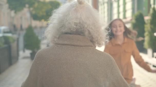 Baksidan av senior man med långt grått hår sitter på urban gata i solljus som två glada barn kör och kramar farfar. Glad vit far- och morförälder som träffar barnbarn utomhus. — Stockvideo