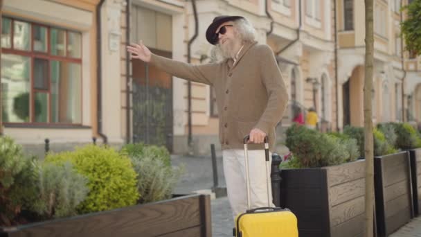 Αυτοπεποίθηση ανώτερος αρσενικό τουρίστας με ταξιδιωτική τσάντα hitchhiking στην πόλη δρόμο. Πορτρέτο του κομψού γέρου Καυκάσου με καπέλο και γυαλιά ηλίου που ταξιδεύει την ηλιόλουστη καλοκαιρινή μέρα. Καλή συνταξιοδότηση έννοια. — Αρχείο Βίντεο