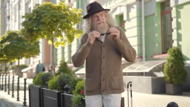 ヨーロッパの街の通りでサングラスやヒッチハイクをする自信に満ちた老人の肖像画。晴れた夏の日にエレガントな白人観光客の退職者の旅行。自信と観光のコンセプト. — ストック動画