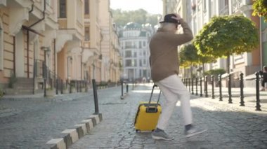 Neşeli yaşlı turist, güneşli bir yaz gününde şehir caddesinde dans edip zıplıyor. Sarı seyahat çantasıyla gezen, kendine güvenen beyaz bir adamın geniş açılı portresi..