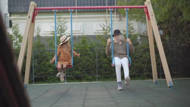 Plan large de grand-père et petite-fille élégants balançant sur des balançoires à l'extérieur. Portrait d'homme âgé caucasien heureux et fille gaie mignonne s'amuser sur la journée ensoleillée en plein air. — Video