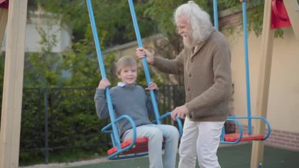 Homem sênior empurrando balanços com o menino sorrindo bonito sentado nele. Retrato de avô caucasiano alegre com cabelos longos cinzentos desfrutando de lazer de fim de semana com neto no parque infantil. — Vídeo de Stock