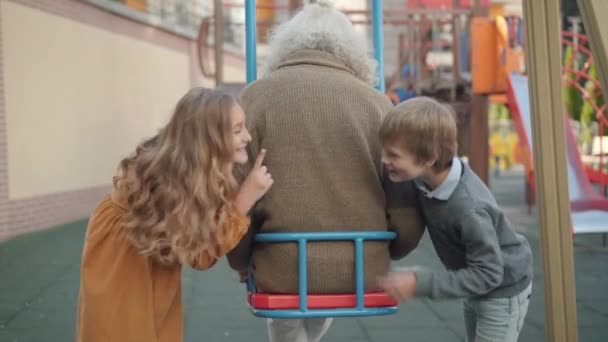 Back view of relaxed old man with long grey hair sitting in swings as cheerful little boy and girl pushing him. Joyful Caucasian grandchildren having fun with grandfather on playground. — Stock Video