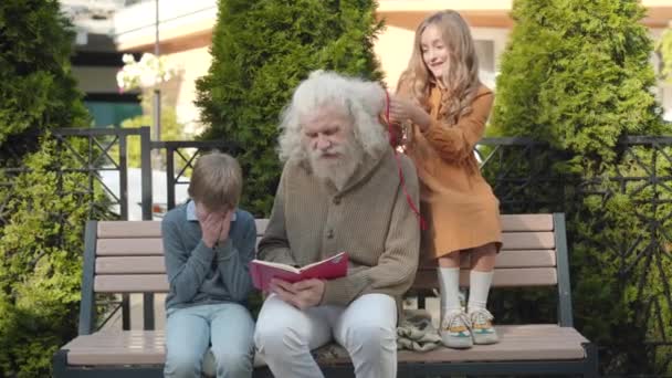 Pretty Caucasian girl making ponytails on long grey hair of grandfather while senior man reading book for smiling little boy sitting on bench. Happy weekend leisure of multi-generational family. — Stock Video