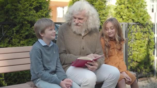 Avô positivo sentado com netos no banco e livro de leitura. Retrato de feliz relaxado caucasiano velho homem, menina e menino desfrutando de fim de semana ensolarado ao ar livre. — Vídeo de Stock