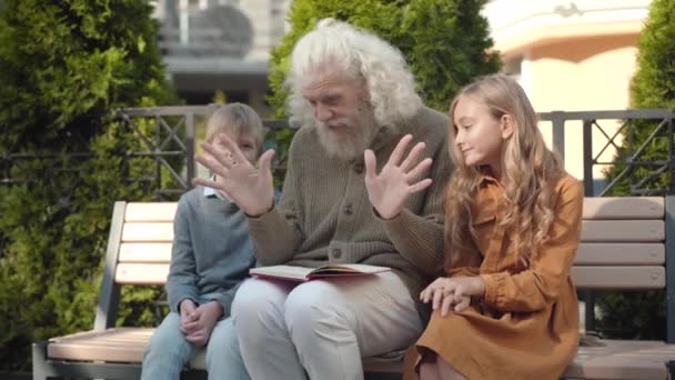 Porträtt av upphetsad farfar gestikulera och prata med glada barnbarn utomhus. Glad intelligent senior vit man diskuterar bok med söt flicka och söt pojke och skrattar. — Stockvideo