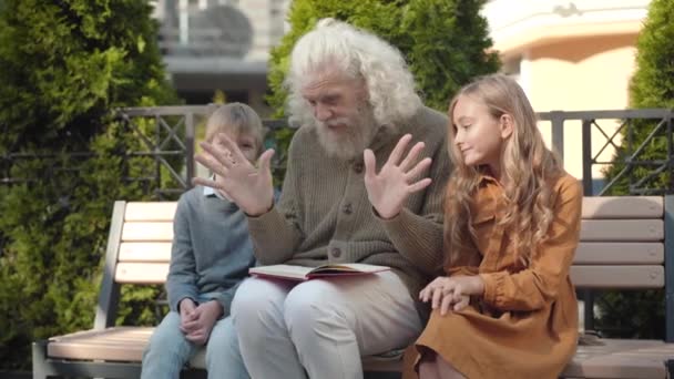 Retrato de abuelo emocionado haciendo gestos y hablando con nietos alegres al aire libre. feliz inteligente mayor caucásico hombre discutir libro con bonita chica y lindo chico y riendo. — Vídeo de stock