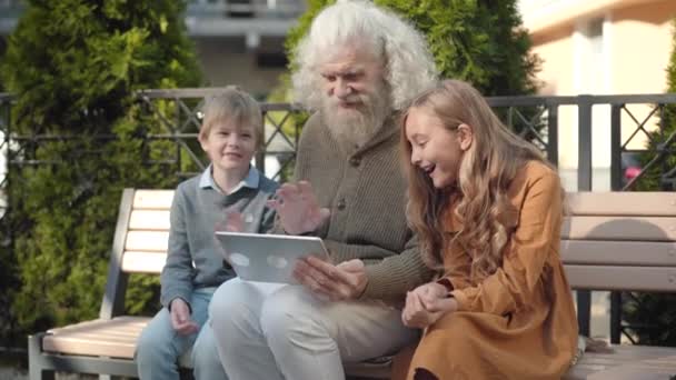 Σύγχρονη Καυκάσιος παππούς κάθεται με τα εγγόνια για ηλιόλουστη μέρα σε εξωτερικούς χώρους και χρησιμοποιώντας tablet με video chat. Χαρούμενος ηλικιωμένος που απολαμβάνει τα σαββατοκύριακα με αγόρι και κορίτσι.. — Αρχείο Βίντεο