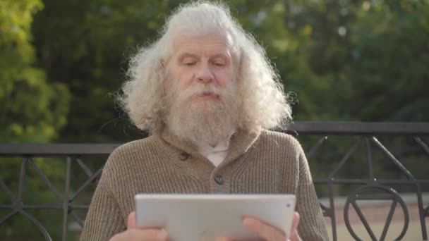 Uzun gri saçlı, güler yüzlü yaşlı sakallı adam gün ışığında dışarıda oturuyor ve tablet selfie makinesine el sallıyor. Açık havada görüntülü sohbet yapan modern Kafkasyalı emeklinin portresi.. — Stok video