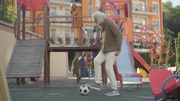 Szerokie ujęcie pozytywnego emeryta grającego piłką nożną na świeżym powietrzu jako dzieci bawiące się na placu zabaw w tle. Portret beztroskiego białego seniora korzystającego z emerytury na świeżym powietrzu. — Wideo stockowe