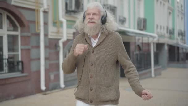 Gondtalan fehér öregember fülhallgatóban, aki a város utcáin táncol. Portré nyugodt boldog nyugdíjas nyugdíjas élvezi napsütéses nap szabadban zenét hallgatni fülhallgató. Szabadidő és életmód. — Stock videók