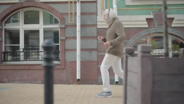 Kameran följer glada roliga manliga pensionär i hörlurar dansar som promenader längs soliga stadsgatan. Sidovy bred bild av glad avslappnad senior man ha kul utomhus med barn passerar. — Stockvideo