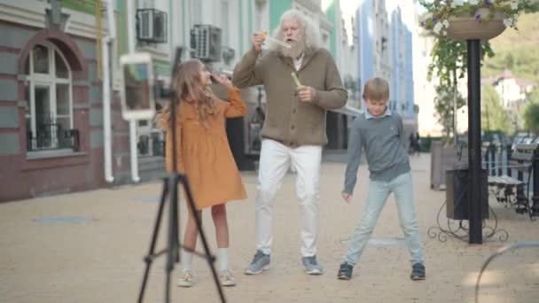 Brede opname van vrolijke kleinkinderen en grootvader die plezier hebben buiten het opnemen van video voor sociale media. Portret van zorgeloze oudere blanke man, kleine jongen en meisje dansen en blazen zeep — Stockvideo