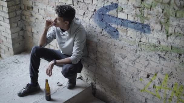 Wide shot of mixed-race teenager smoking and drinking beer on urban ruins in ghetto. Top view portrait of depressed teenage boy having bad habits. Homeless teen on slum. — Stock Video