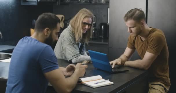 Tři pozitivní univerzitní studenti sedí v hostelu kuchyně a mluví. Portrét veselých mladých mužů surfujících na internetu na smartphonu a notebooku a chatování. Cinema 4k ProRes HQ. — Stock video