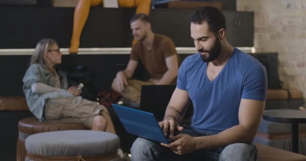 Confident Middle Eastern man working online on laptop in casual coworking as friends talking at the background. Πορτρέτο του όμορφου νεαρού που γυρίζει στην κάμερα και χαμογελάει. Κινηματογράφος 4k ProRes HQ. — Αρχείο Βίντεο