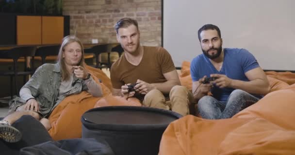 즐거운 천년의 동료들이 공동의 공간에서 휴식 시간에 게임 콘솔을 가지고 놀고 있습니다. 편안 한 젊은 남자들이 자유 시간을 즐기고 있다. 시네마 4k ProRes HQ. — 비디오