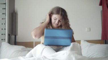 Sabahları yatakta oturup dizüstü bilgisayar kullanan konsantre genç bir kadın. Karantina sırasında internette çalışan tombul beyaz kadının portresi. Uzaktan çalışma ve serbest çalışma.