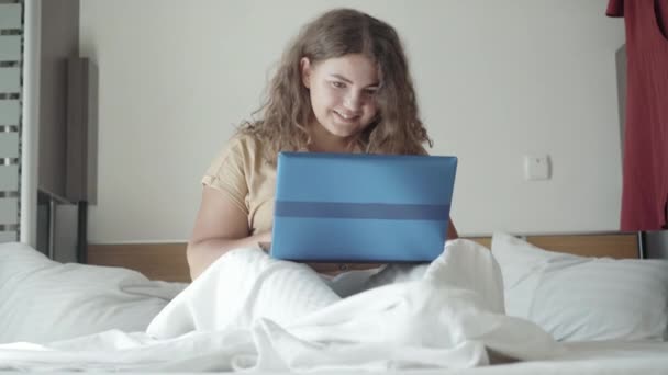 Portrait of happy charming plus-size woman using social media on laptop sitting in bed in the morning. Young chubby Caucasian lady messaging online in bedroom. Device addiction concept. — Stock Video